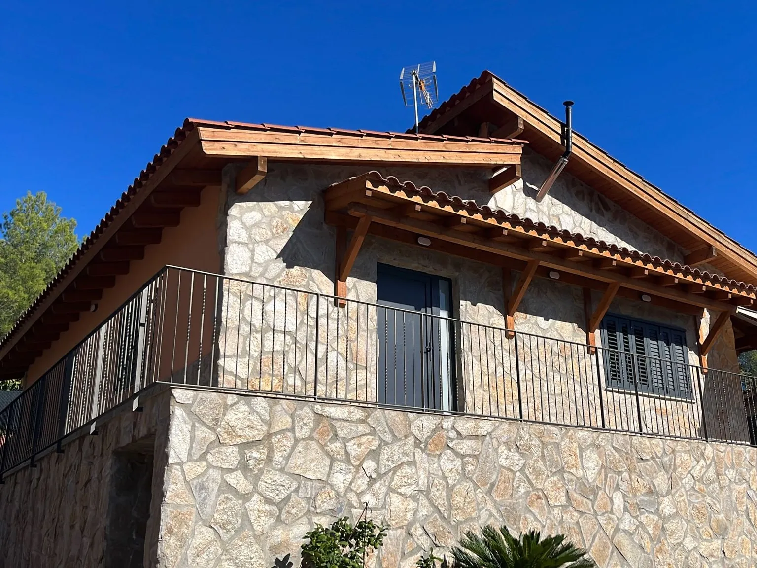 Casa con la fachada y el muro revestidos de piedra, tejado de teja con aleros de madera y barandilla negra